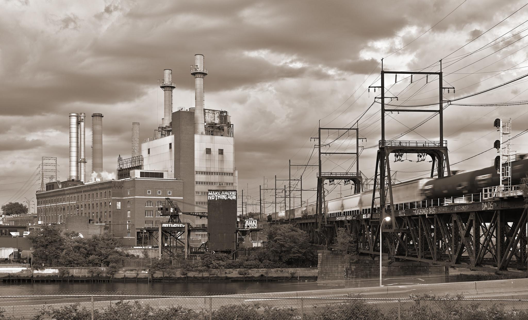 A mid-century power plant, with four prominent smokestacks atop an industrial building, at left across a small river. At right, a railroad drawbridge crosses the river, with a freight train slowly streaking across.