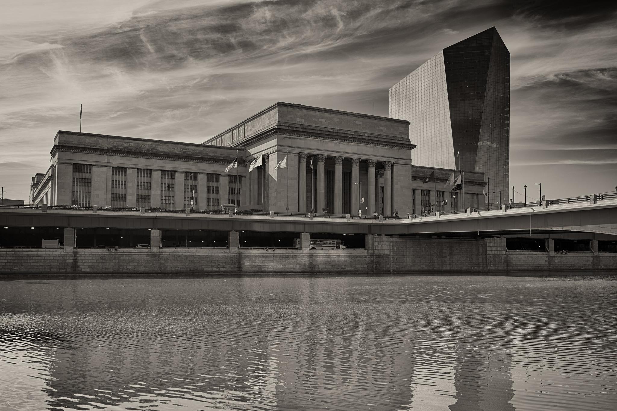 A large neoclassical train station across a river. To the right of and behind the station is an irregularly-shaped modernist glass office tower.