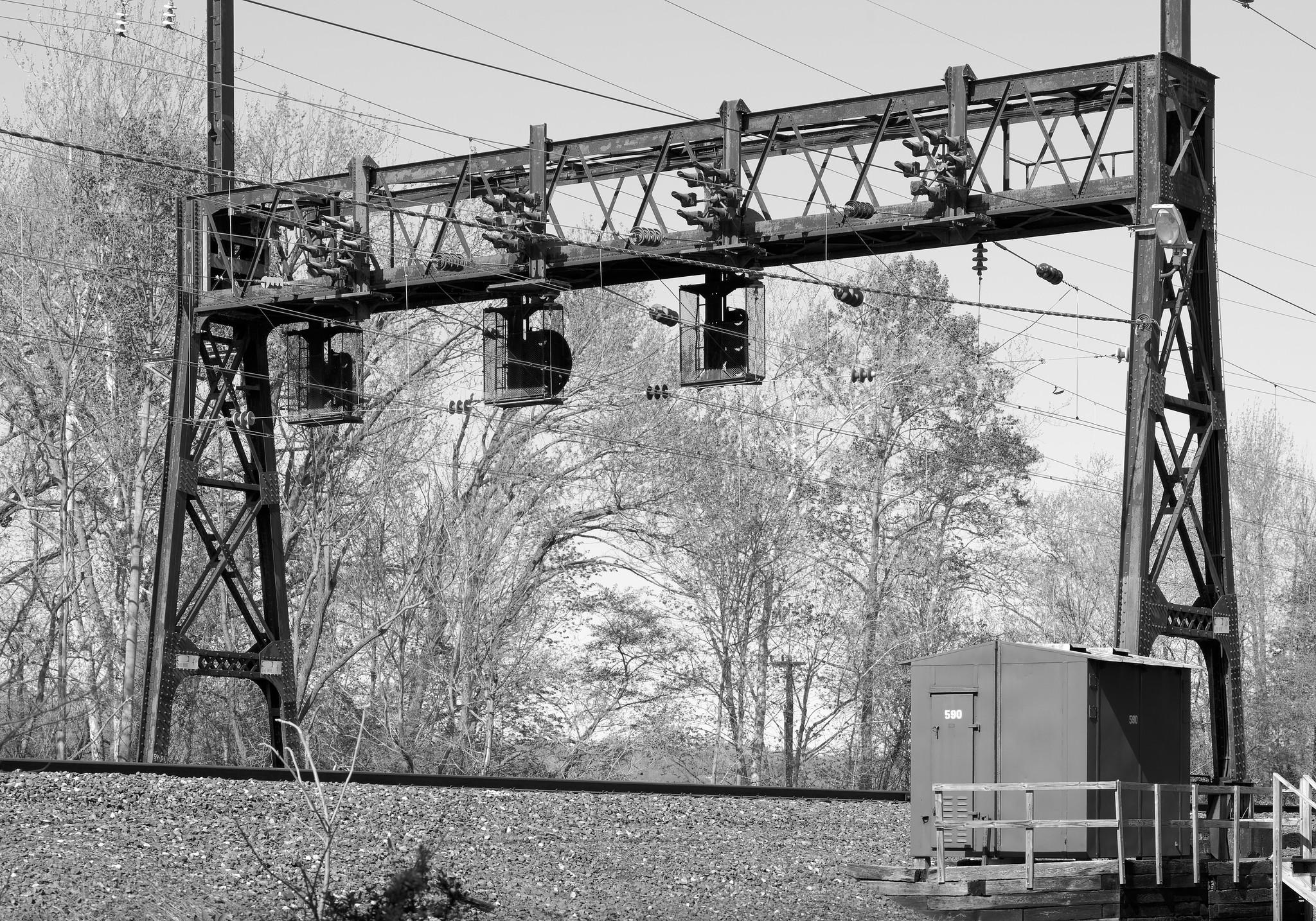 A steel "signal bridge" over a railroad line, supporting high voltage overhead catenary wires and with illuminated signals above each of the four tracks.