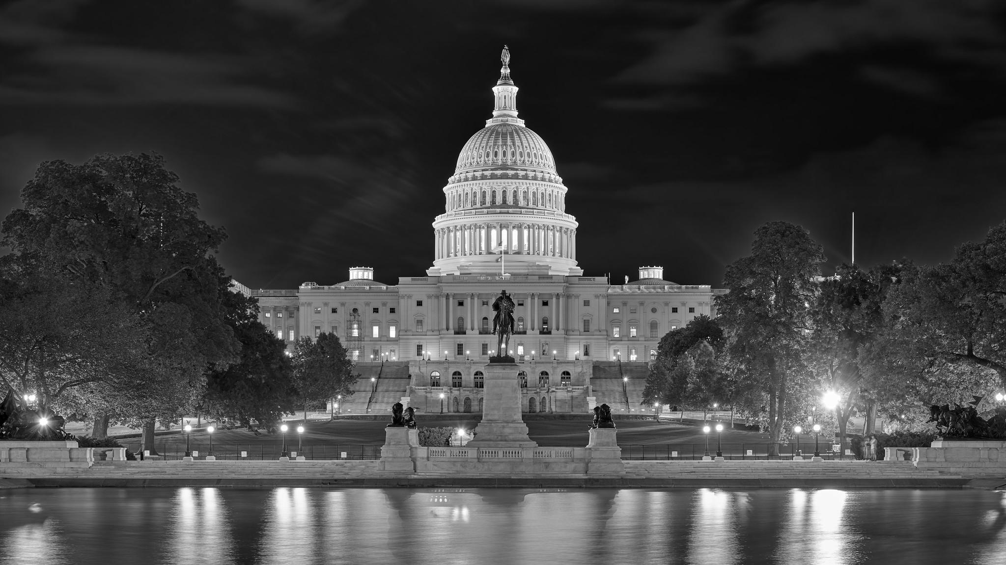 The western side of the US Capitol building at night, with its prominent dome illuminated. Reflections are visible in the pool at foreground.