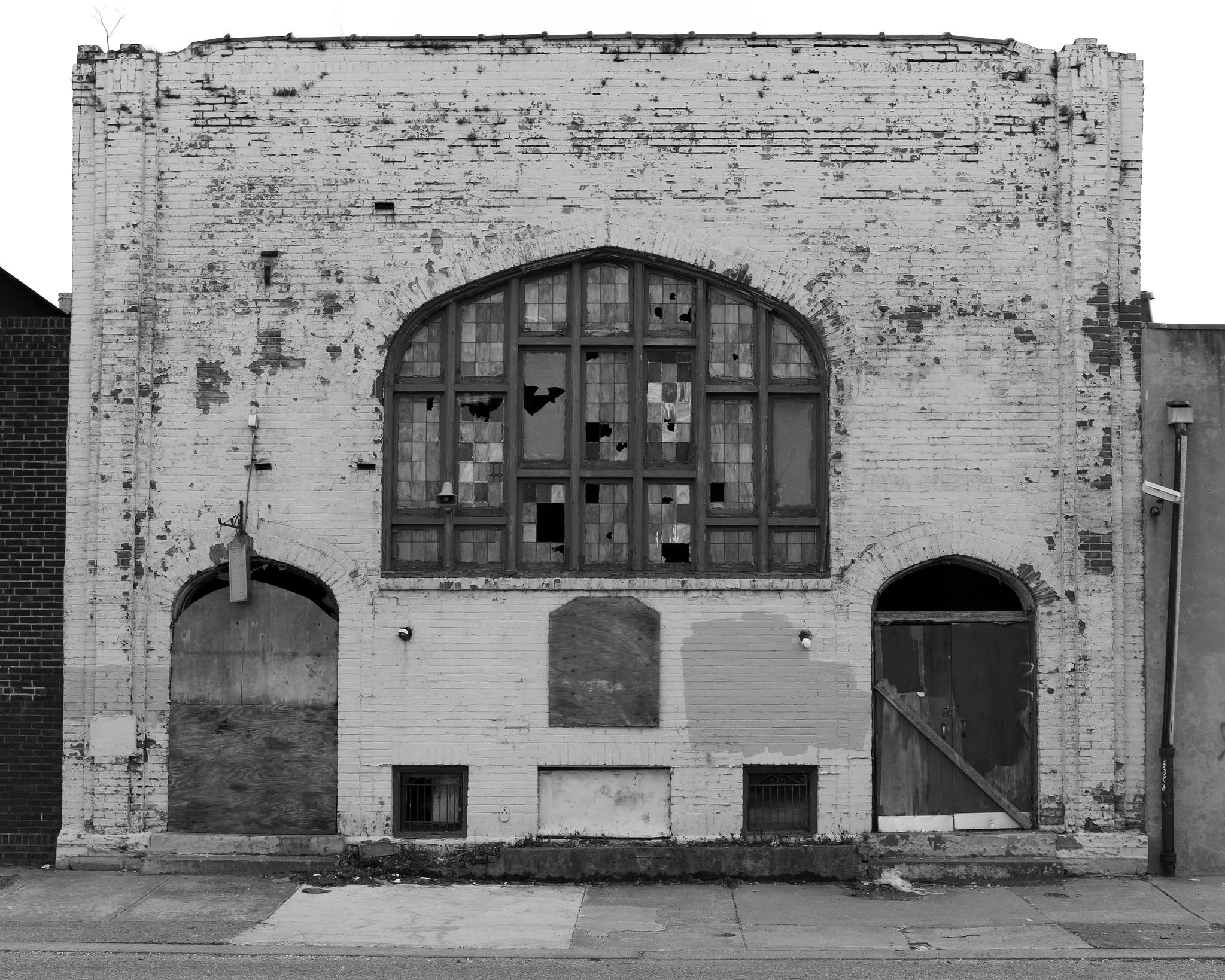 A abandoned, derelict urban church, with a simple rectangular facade and a large broken window at center.