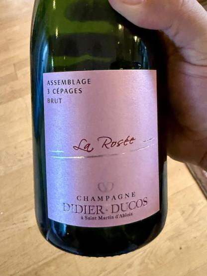 A close-up photograph shows a hand holding a dark green glass bottle against a wooden floor background. The bottle features a light pink label with black and red text. At the top, the label reads "ASSEMBLAGE", "3 CÉPAG CÉPAGES", and "BRUT". In the center, "La Rosée" is written in a reddish cursive font between two thin horizontal lines. The bottom of the label contains a small logo above the words "CHAMPAGNE", "DIDIER - DUCOS", and "à "à Saint Martin d'Ablois".

🌱 Energy used: 0.190 Wh