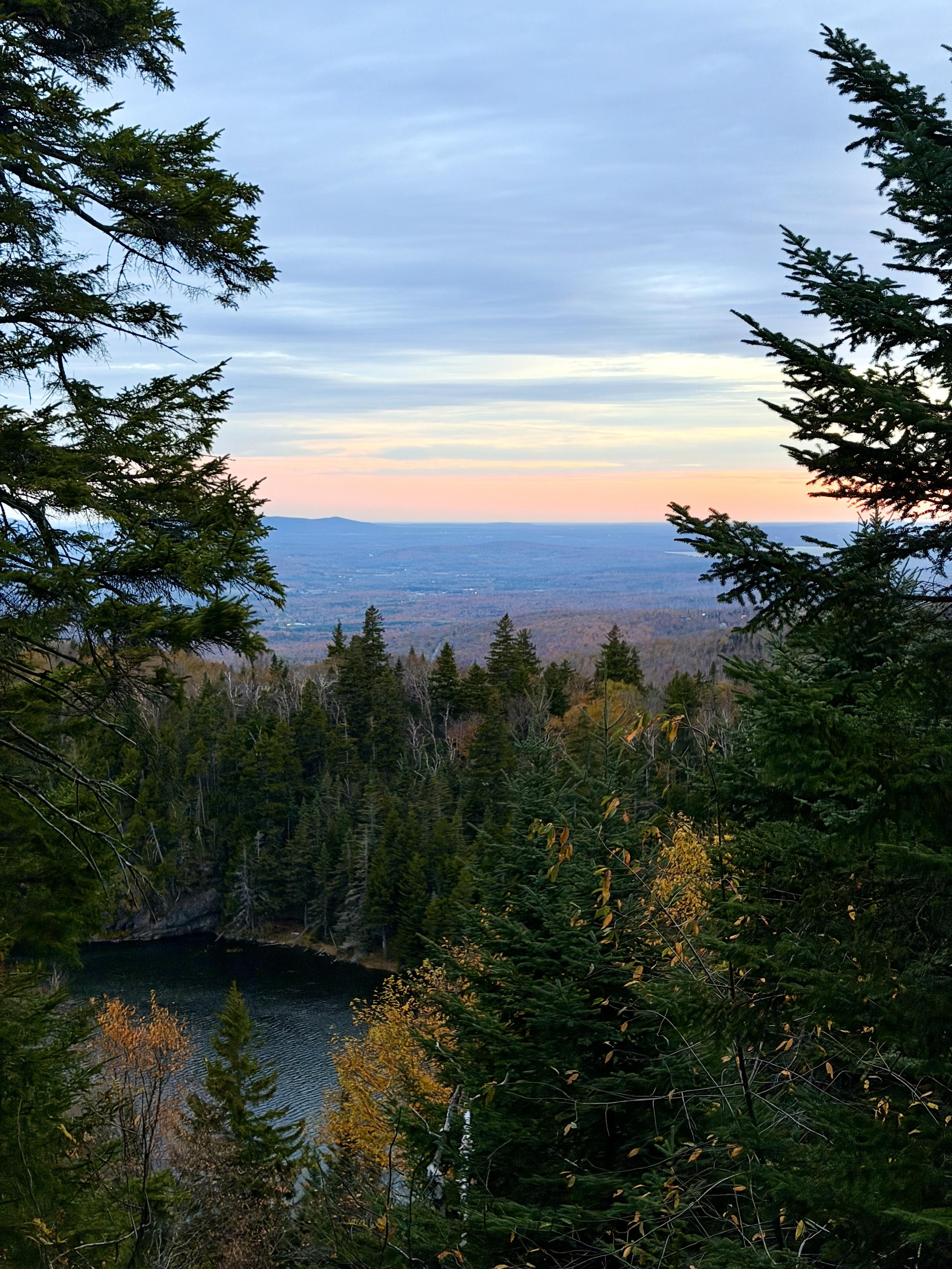 Des couleurs de lever de soleil au fond, des couleurs d’arbres à l’automne au devant et un lac en contre bas.