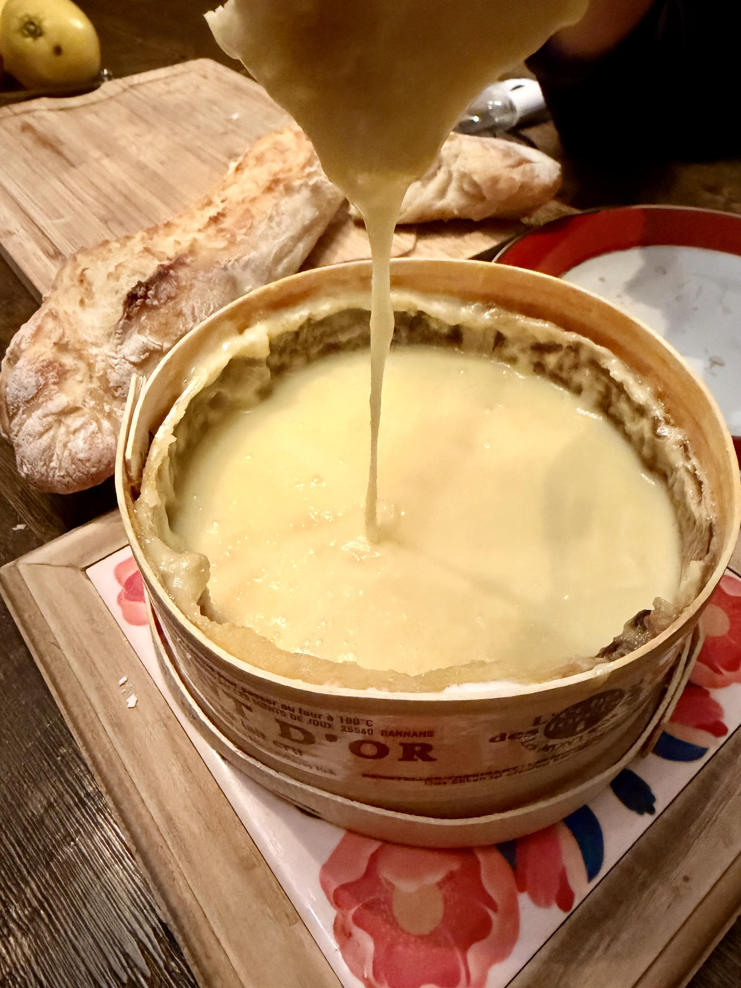 Un fromage Mont d’or fondu avec un bout de pain qui dégouline de fromage dedans (on voit le fil), en arrière plan une baguette au levain maison.