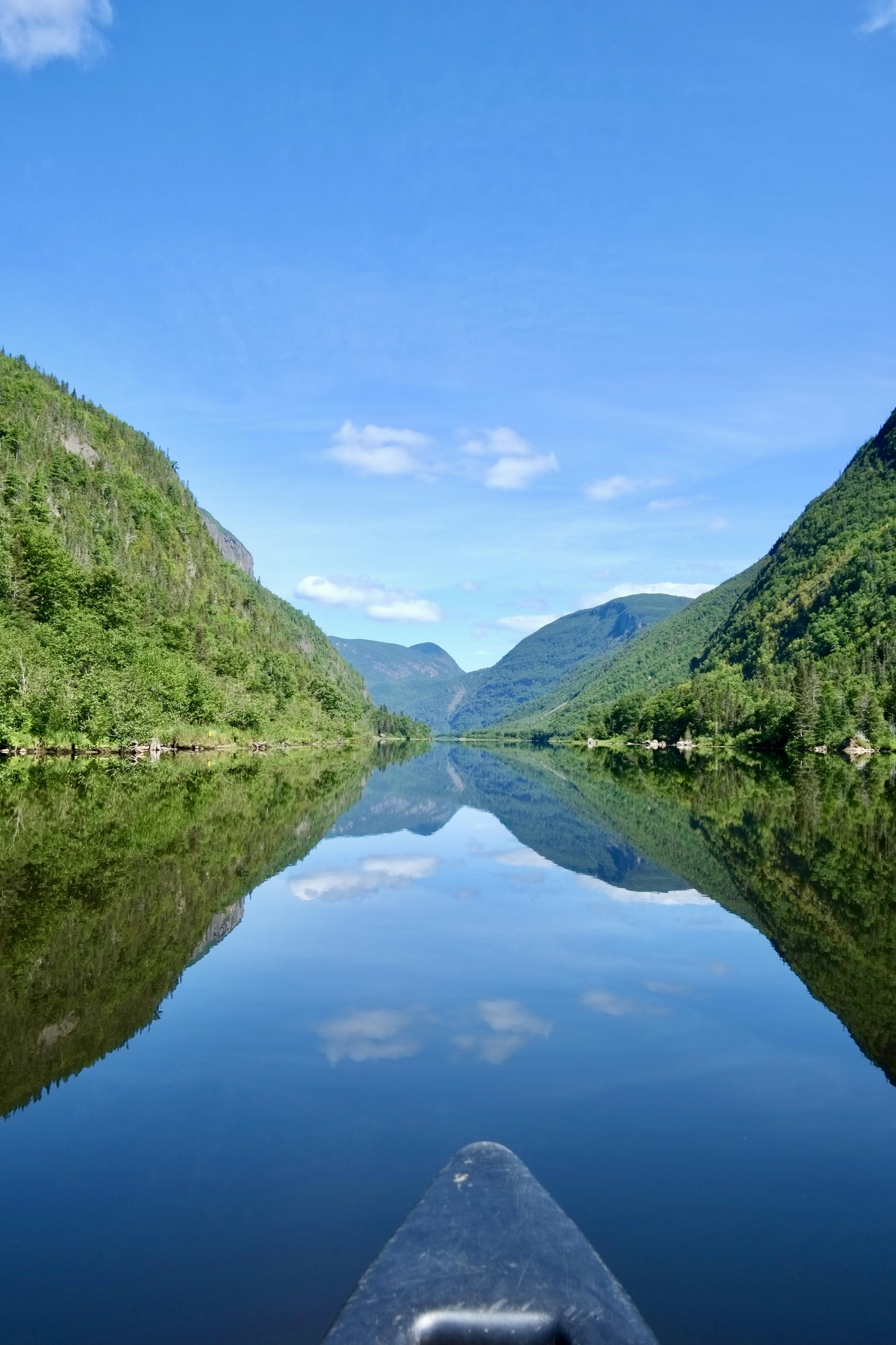 Photo prise depuis un canot au milieu d’un lac avec un reflet parfait des collines environnantes. On voit la pointe du canot.