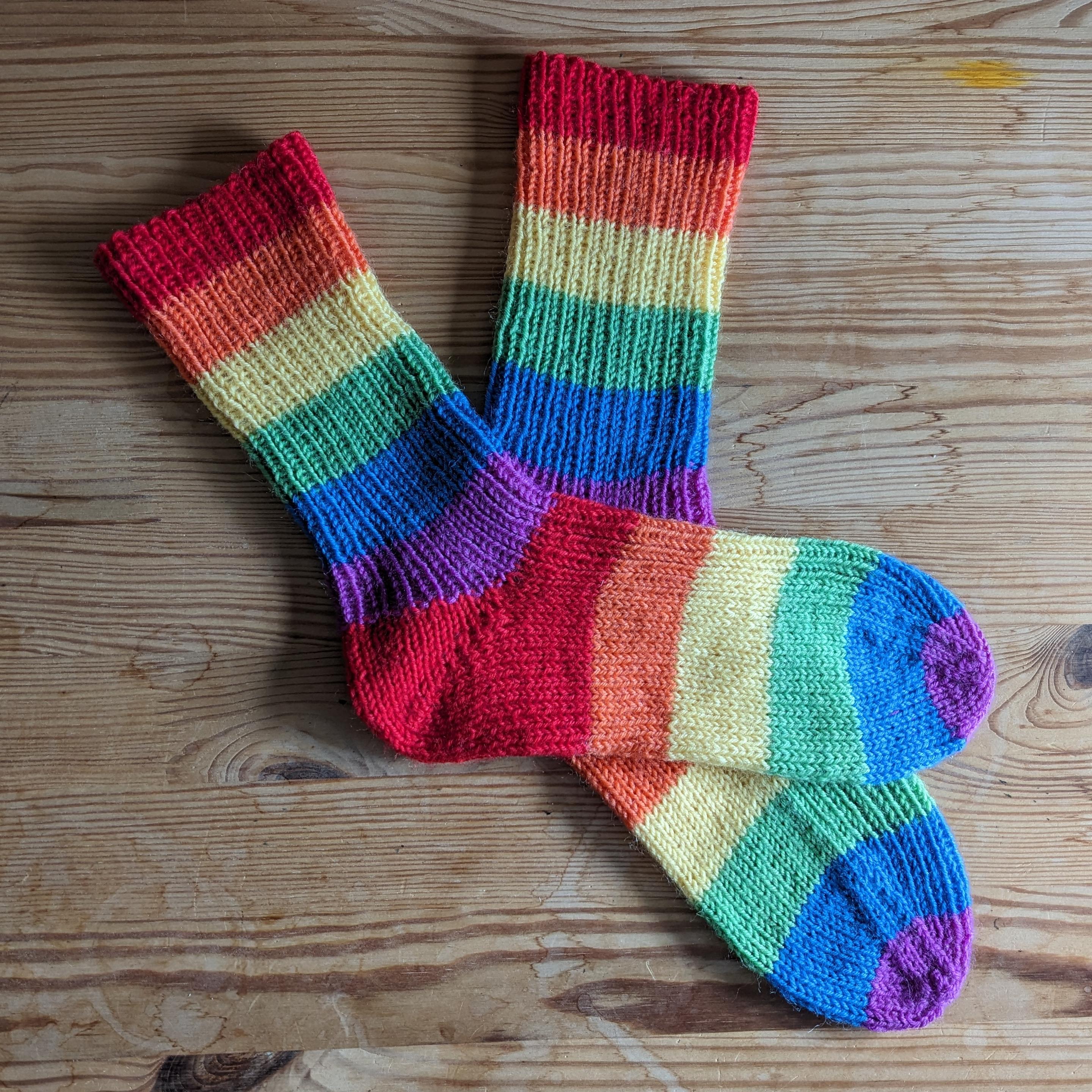 Pair of pride flag socks. Wide stripes from toe to cuff: purple, blue, green, yellow, orange, red, heel, purple, blue, green, yellow, orange, red.