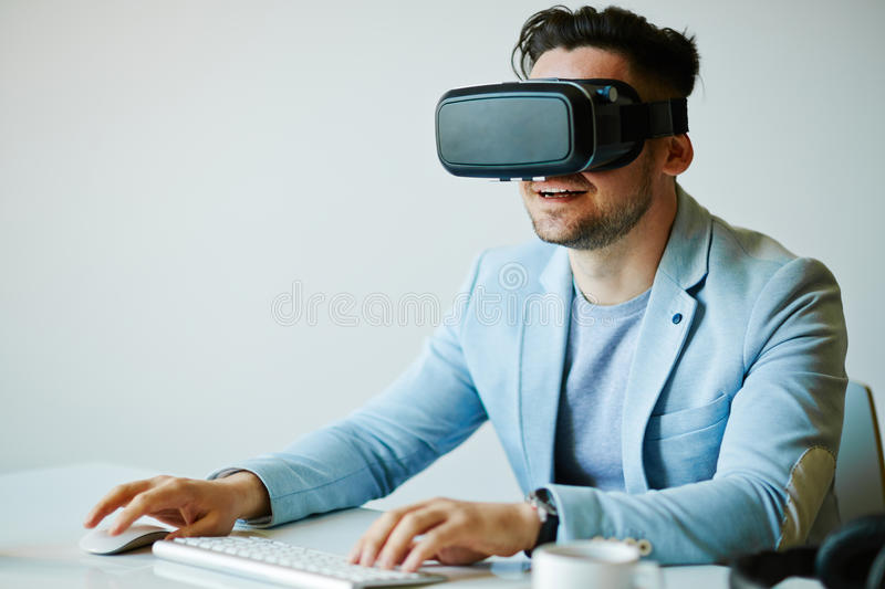 Man in a light blue suit sits at a table with VR goggles on, holding a mouse and a keyboard