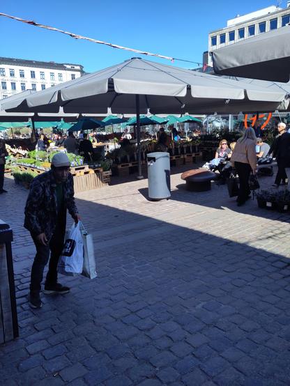 Man ser et blomstermarked med mennesker der sidder i solen ved borde.