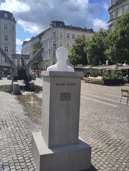Bust af Elon Musk på en solbeskinnet plads i en by.