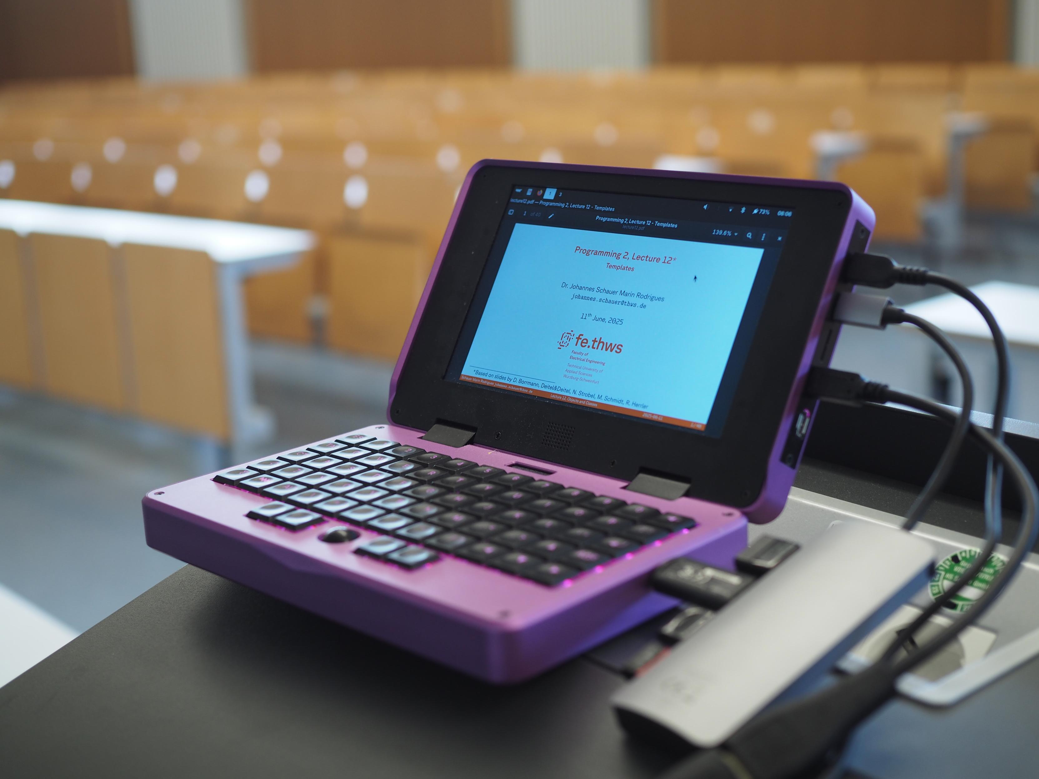 A purple MNT Pocket Reform mini laptop in an empty lecture hall, showing lecture slides on its screen. It is charging via USB-C, is connected to the projector via HDMI and has a few USB devices connected via a USB-C hub.