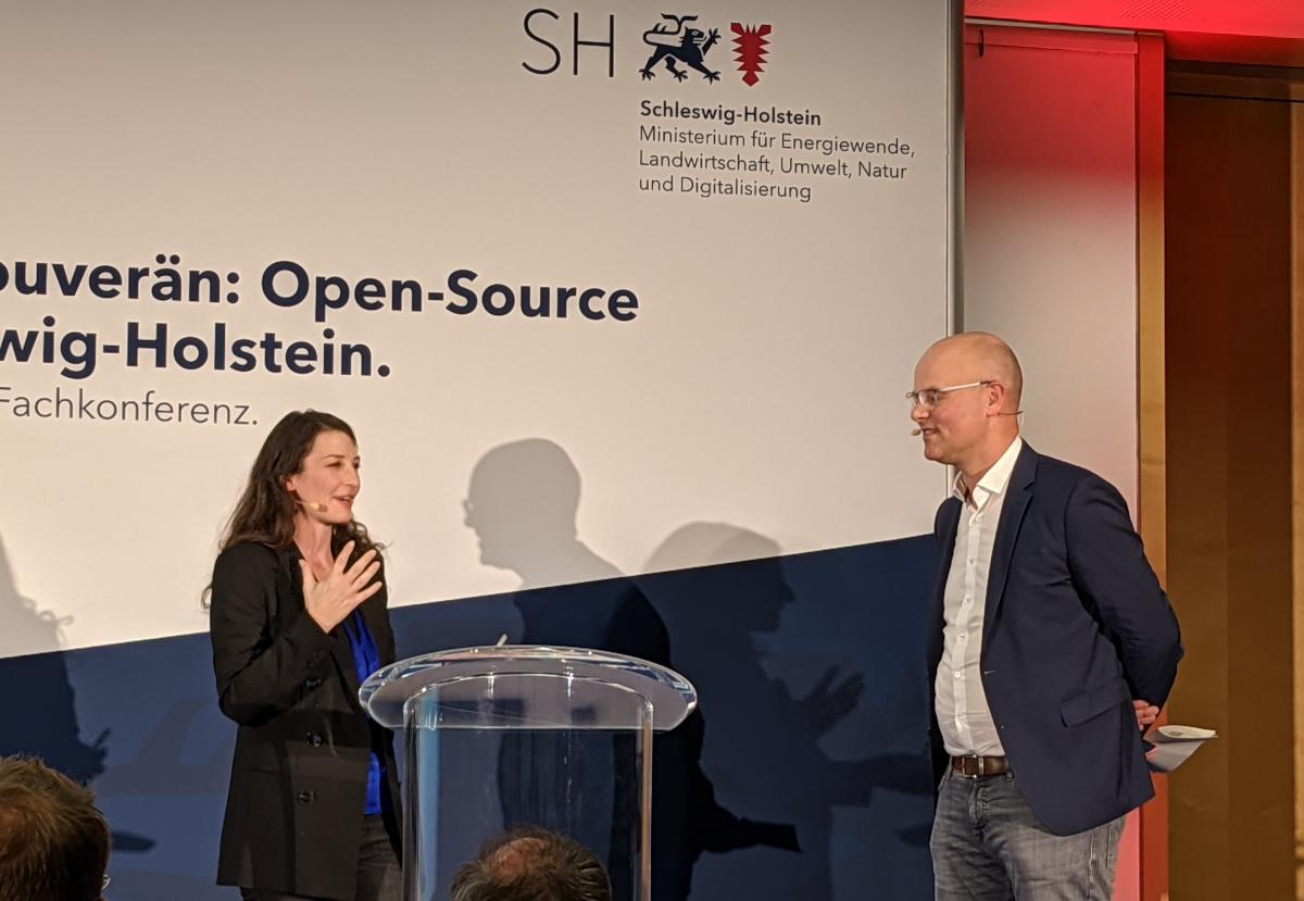 Two people take the stage at a conference. Behind them we can read "Schleswig-Holstein" and "Open-Source" on the slide.