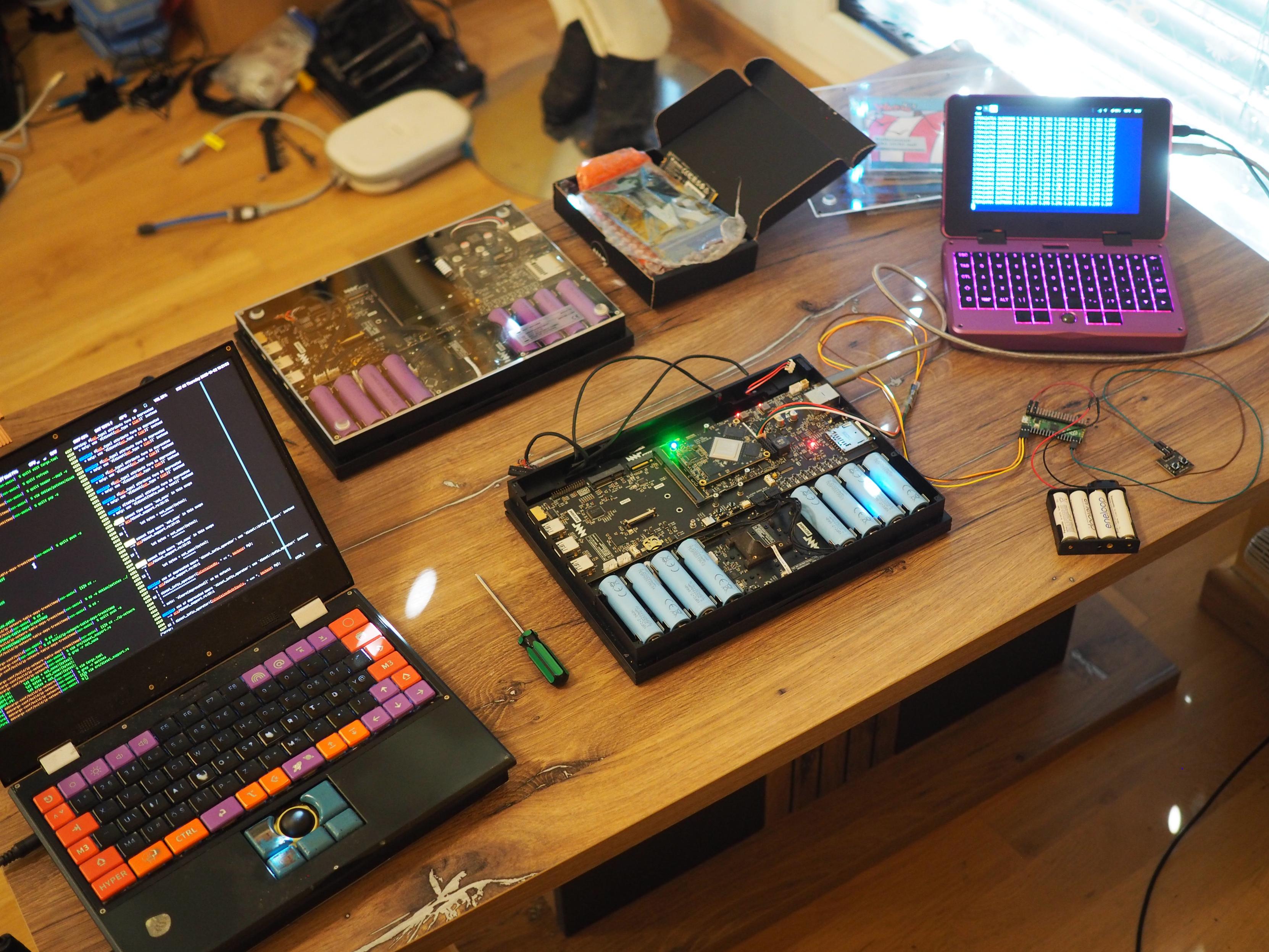 A small table top with four laptops on it. On the far left, the MNT Reform with BananaPI A311D. In the center two classic Reform upside down. The closer of the two has its transparent acrylic bottom removed so that one can see the Rockchip RK3588 and blue battery cells. The other has purple battery cells and contains the i.MX8MQ SoM. Next to it is a black open cardboard box with the LS1028A SoM in it. On the far right is a purple MNT Pocket Reform connected to the RK3588 Reform via a USB-C cable. Its screen shows the serial terminal output from the Rk3588 classic Reform, running a logging script. A small battery pack with four rechargeable AA batteries powers a tiny RP2040 board which is able to send the power-on signal to the RK3588 Reform when a button is pressed.