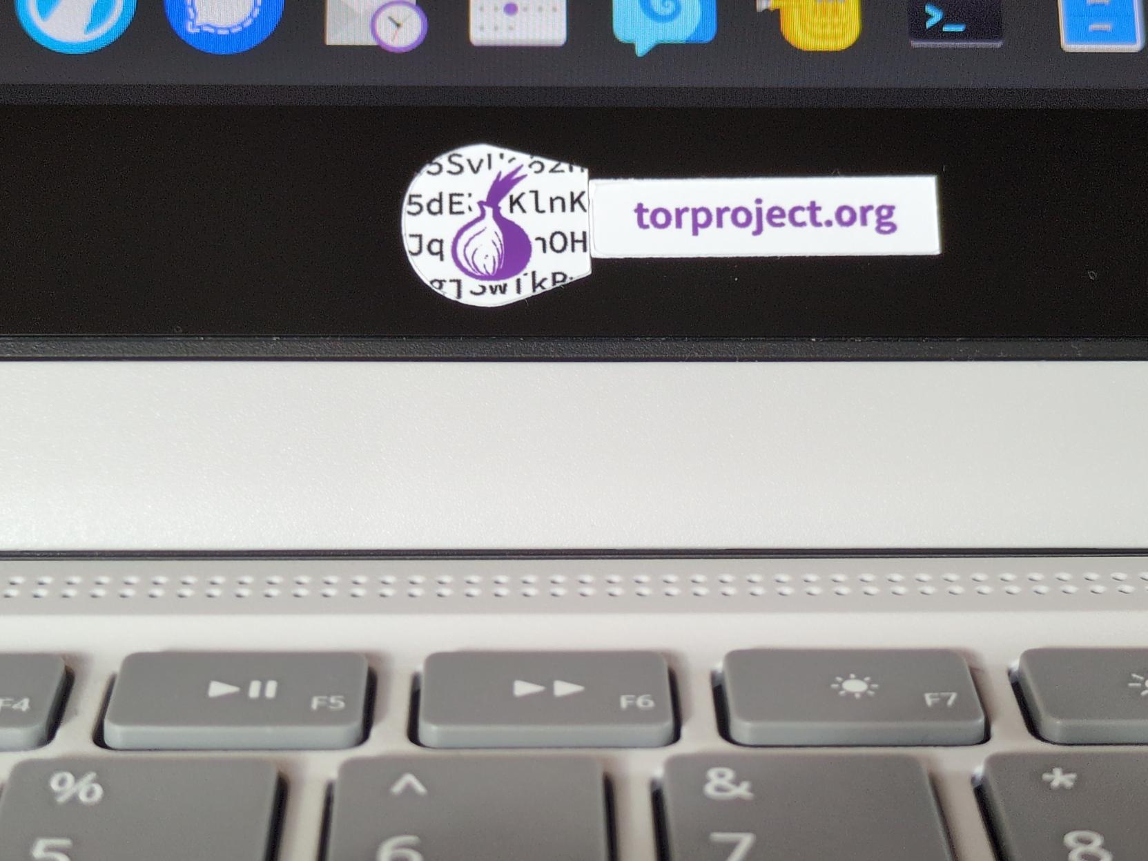 Two small Tor Project stickers at the bottom of the display bezel of a Framework Laptop 12. One round-ish sticker is their onion logo and one has the text "torproject.org" in purple. A partial keyboard and desktop can also be seen.