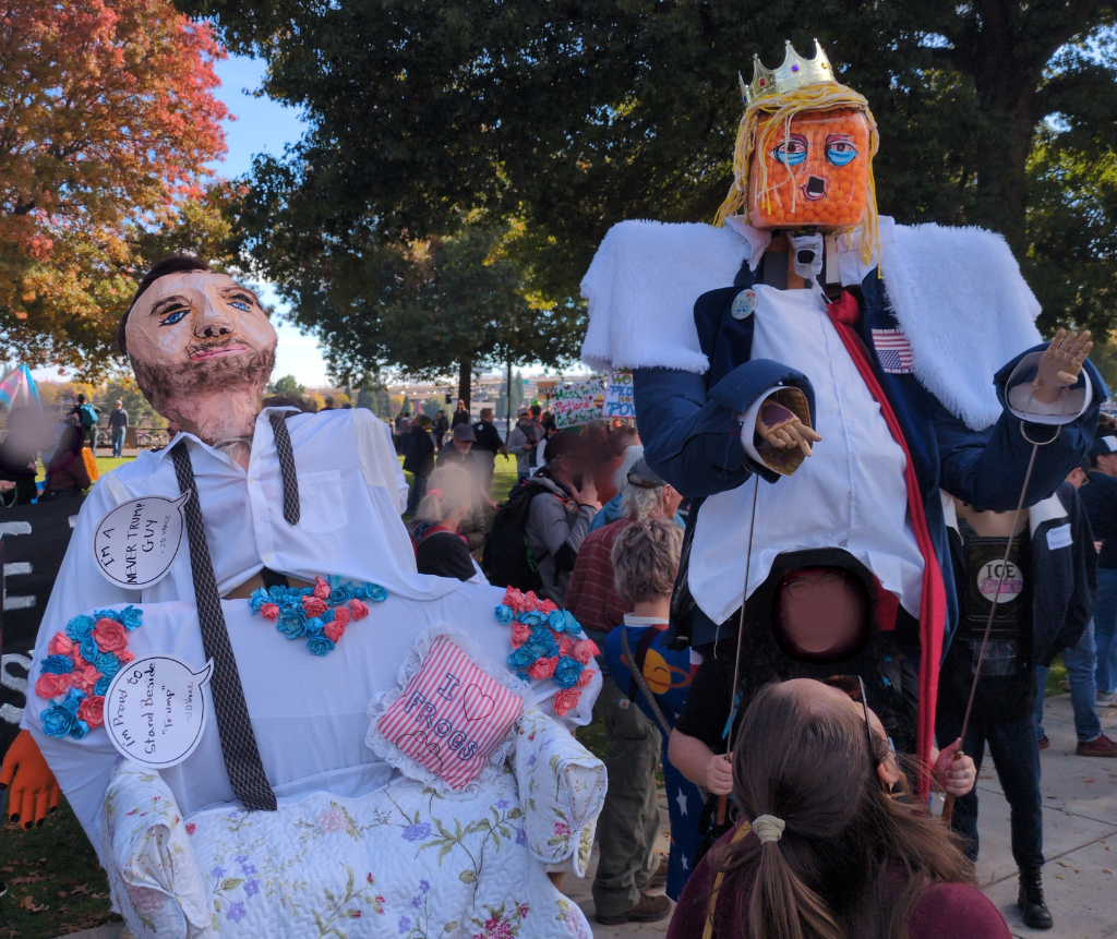 Giant 2x size puppets of Trump and JD Vance. Trump is wearing a crown and his head is made of a cubic plastic container of orange cheez balls.