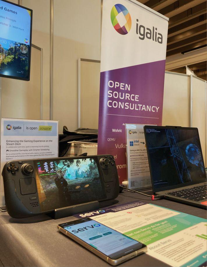 Photo of Igalia's Booth #12 at OSS Japan 2025. There are many devices with different demos: TV, Steam Deck, smartphone and laptop. There are also some boards describing the work done by us on each device.