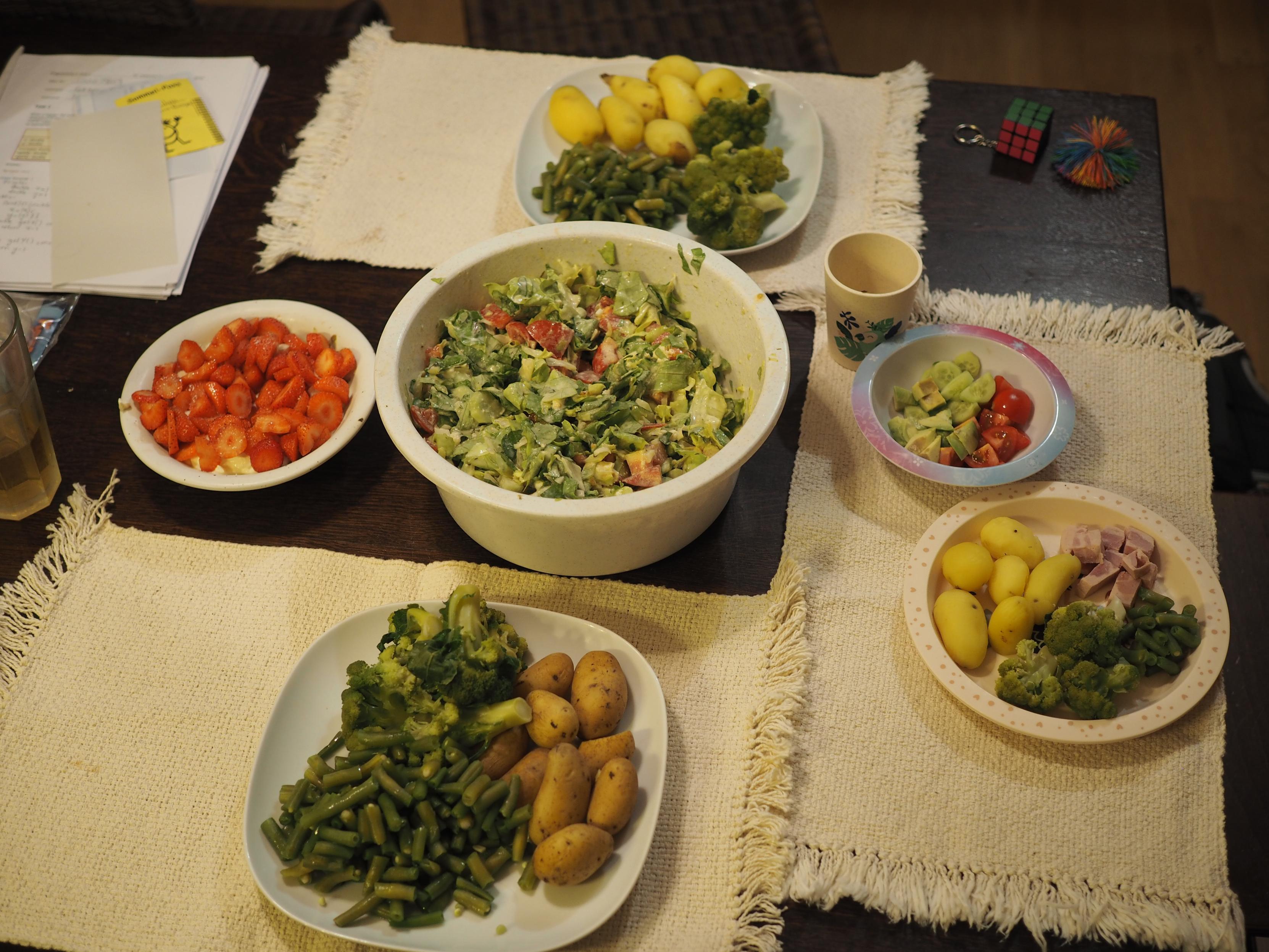 A table set for three people with three plates and a bowl of salad in the middle. The dark table has three white mini-table cloths on it, one under each of the white plates. The plates contain various cooked vegetables like broccoli, potatoes, peans and ham. The salad contains conventional salad leaves, tomato, onion, cucumber and mozzarella in a white dressing based around mayonnaise, vinegar, sunflower oil and leftover liquid from sour pickles. One of the plates has a tiny bowl on the side with cut avocado, tomato and cucumber. On the opposite end is another tiny bowl with strawberries on quark.