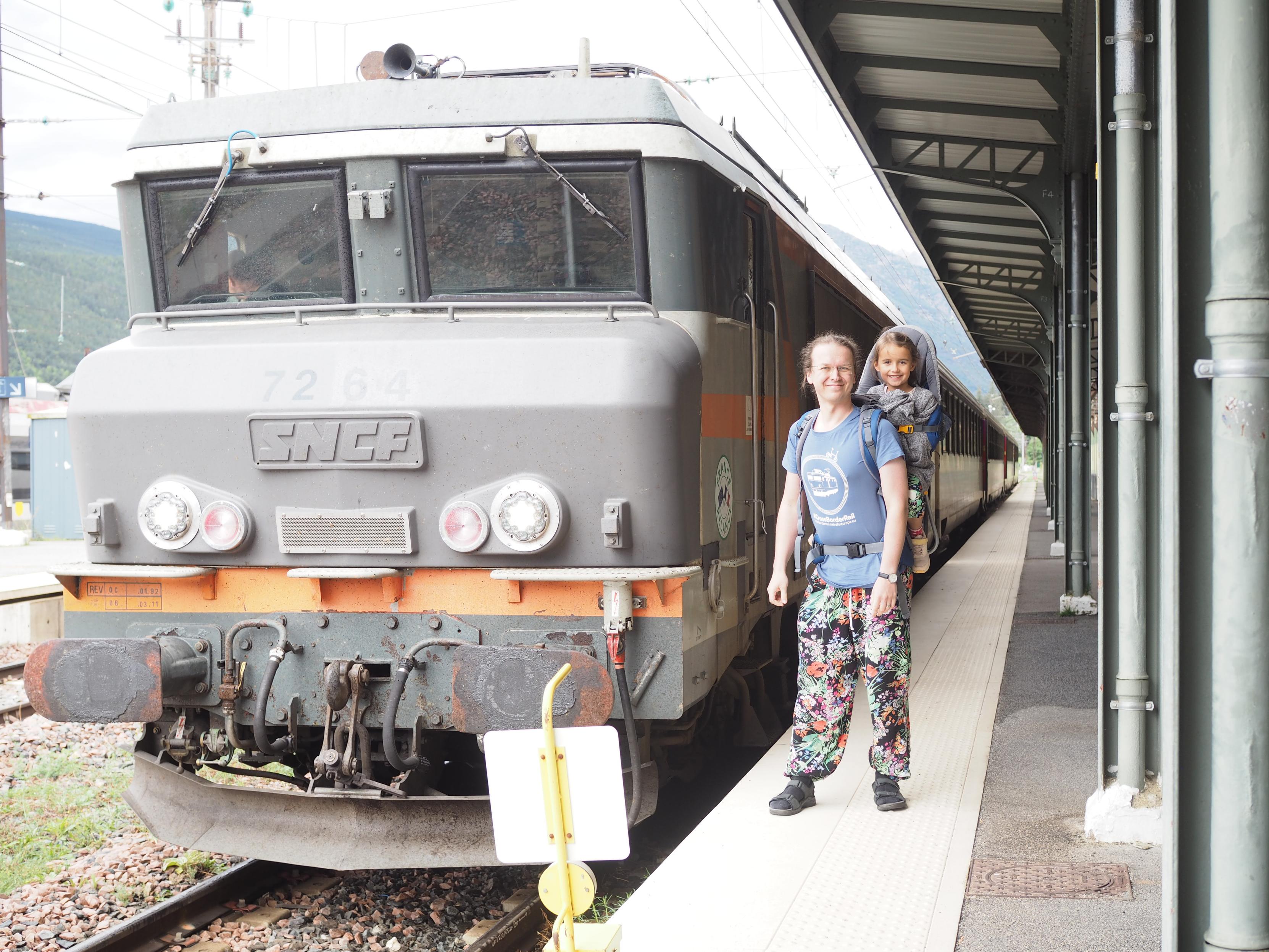 A platform of a train station. An old gray train used for pulling sleeper wagons is waiting on the track. I am standing on the platform facing the camera. A wear a backpack with my daughter in it. I wear colorful flower pants and a blue t-shirt from the #crossborderrail merchandise shop.