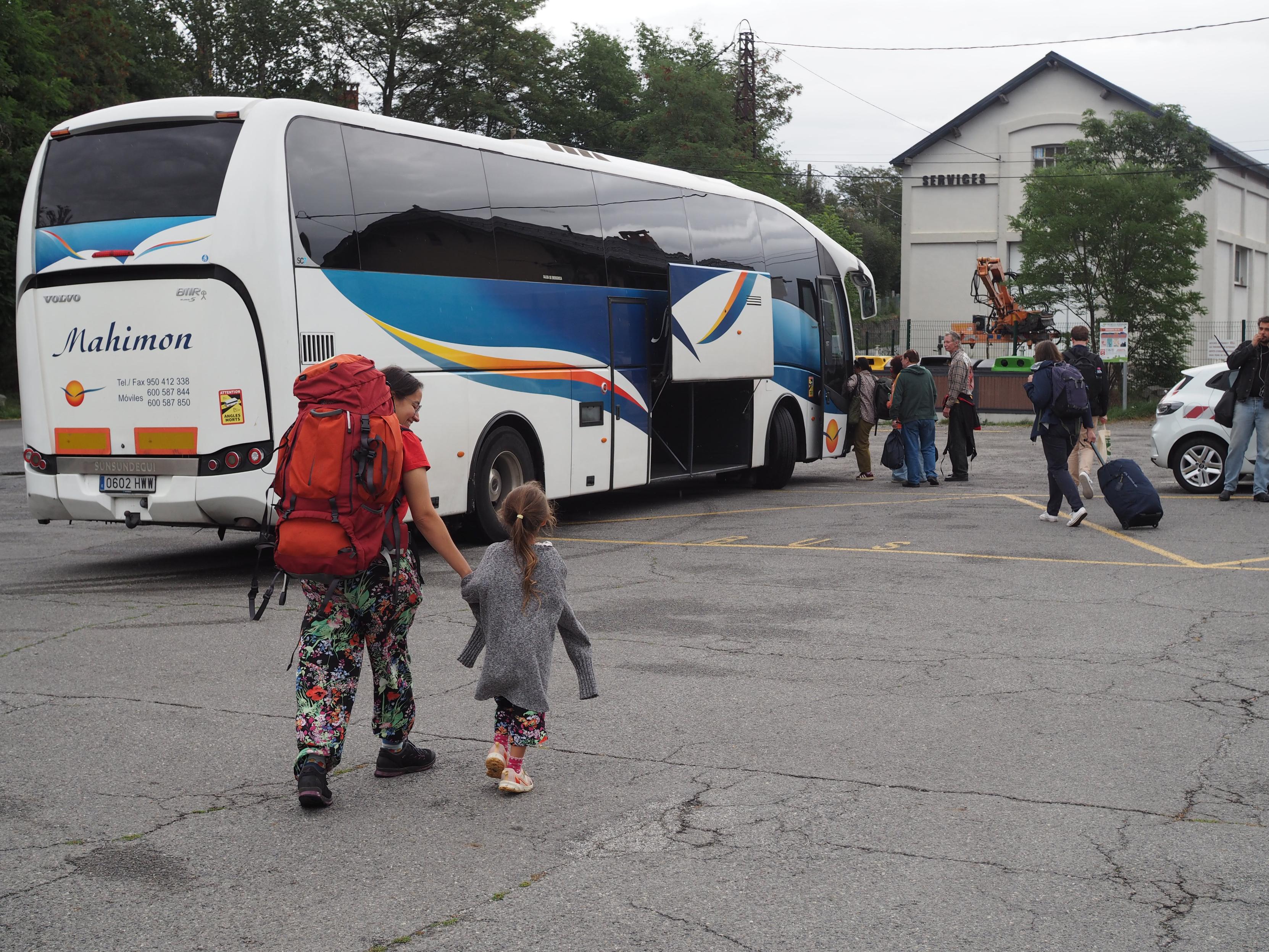 Concrete road with a white bus with a blue design on it in the background. People are walking towards it with a lot of luggage. In the front, my partner and daughter with their own luggage walking towards the bus, photographed from the back.