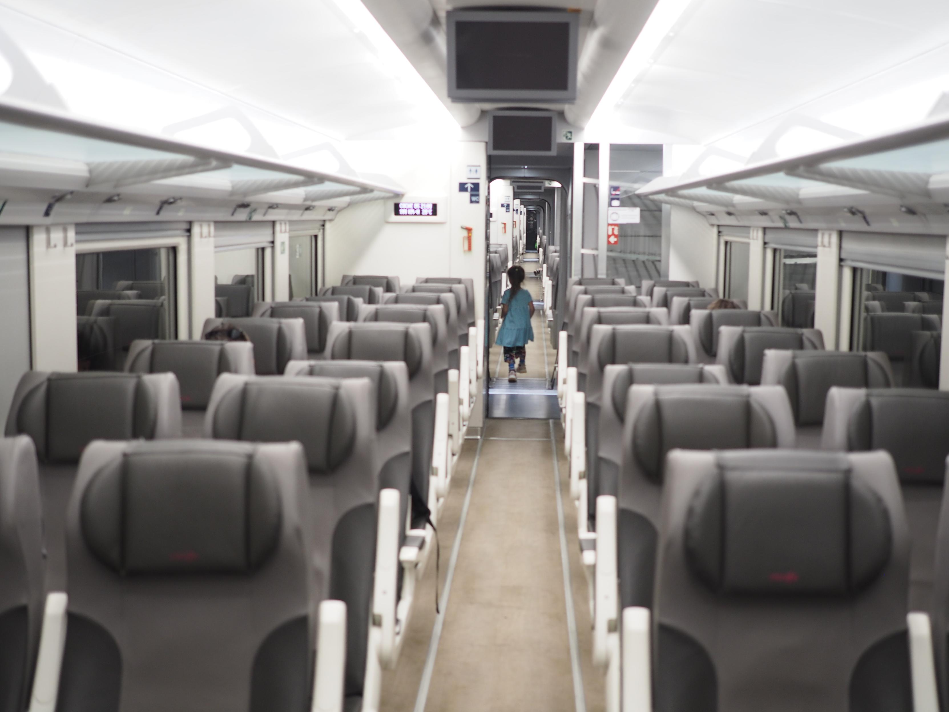 A view down a nearly empty spanish Ave train. A lot of empty gray seats can be seen inside a white-walled carriage. A child in a light blue dress is running down the aisle between the seats. A barely readable display informs that the train is going at 200 km/h and it's 30°C outside.