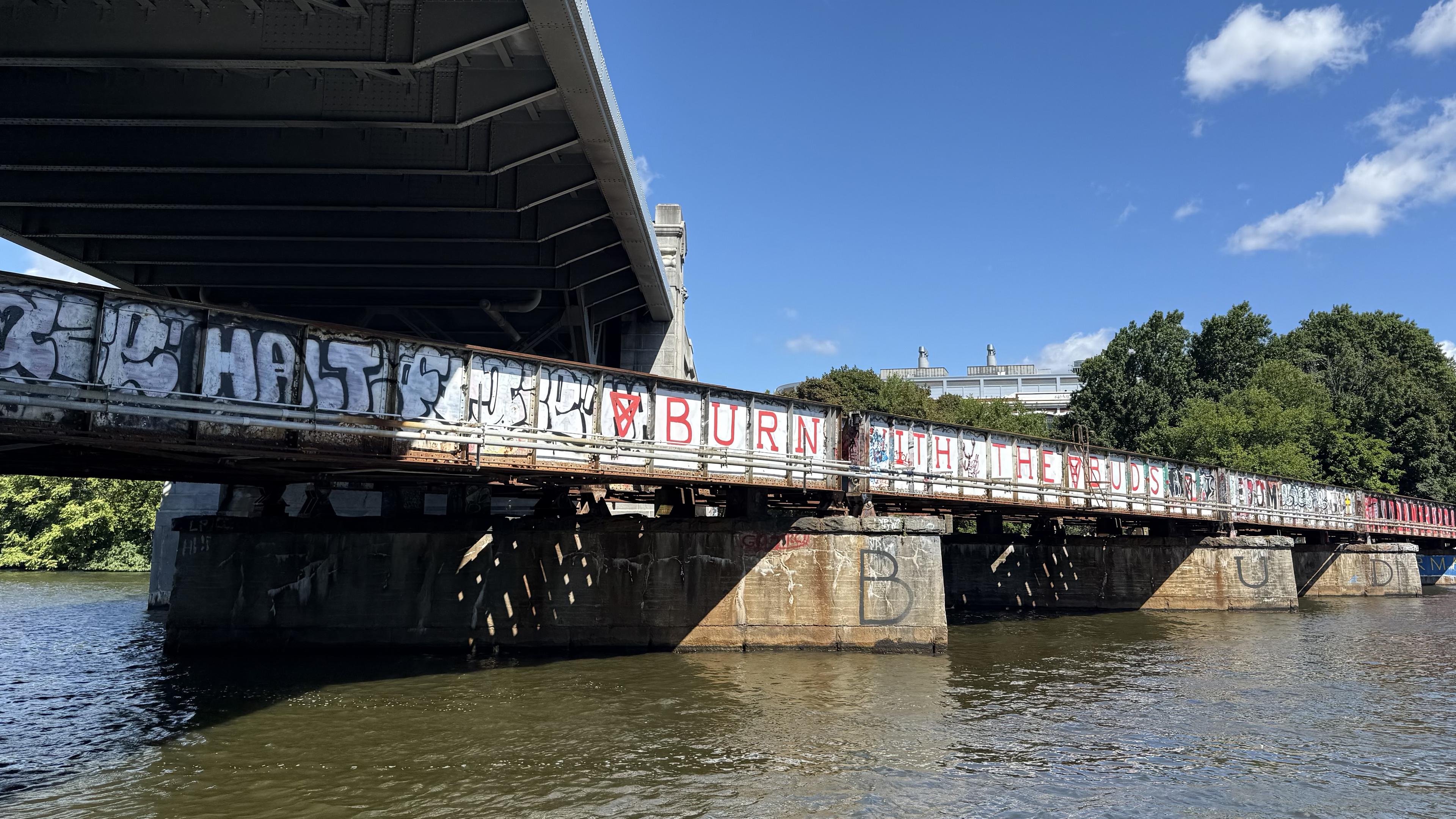 Photograph of a railway bridge across the Charles River. Above it runs a much taller and wider bridge for cars and pedestrians. The lower bridge is covered with graffiti.