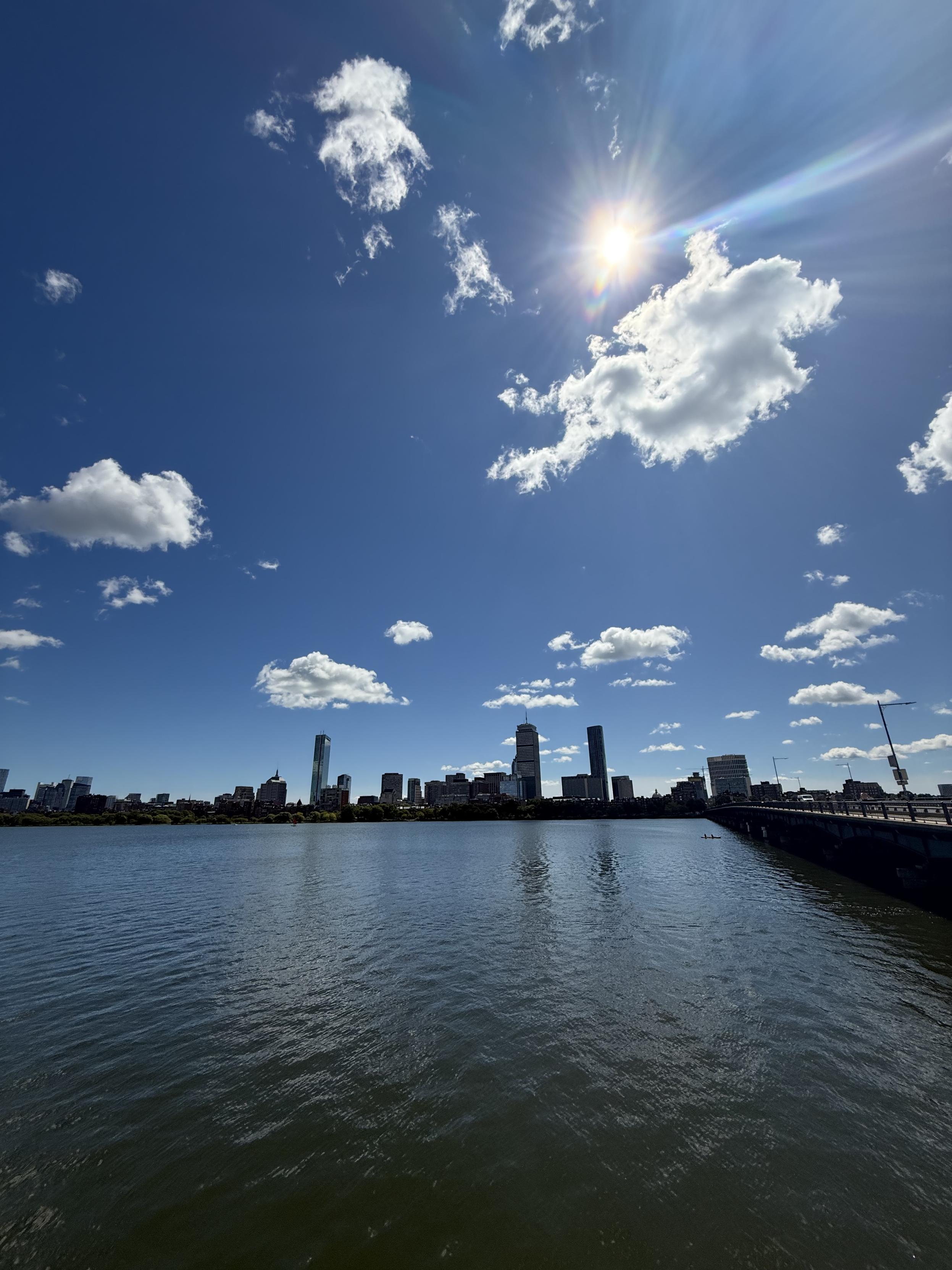 A zoomed-out photograph of the Charles River underneath a sunny, cloud-flecked sky. The Boston skyline dots the river’s far shore.