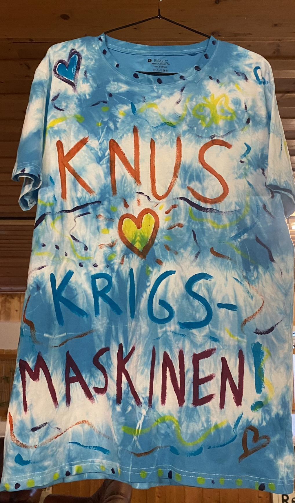 A t-shirt with fresh trippy patterns in shades of blue, green and red. It carries the text "knus krigsmaskinen!"