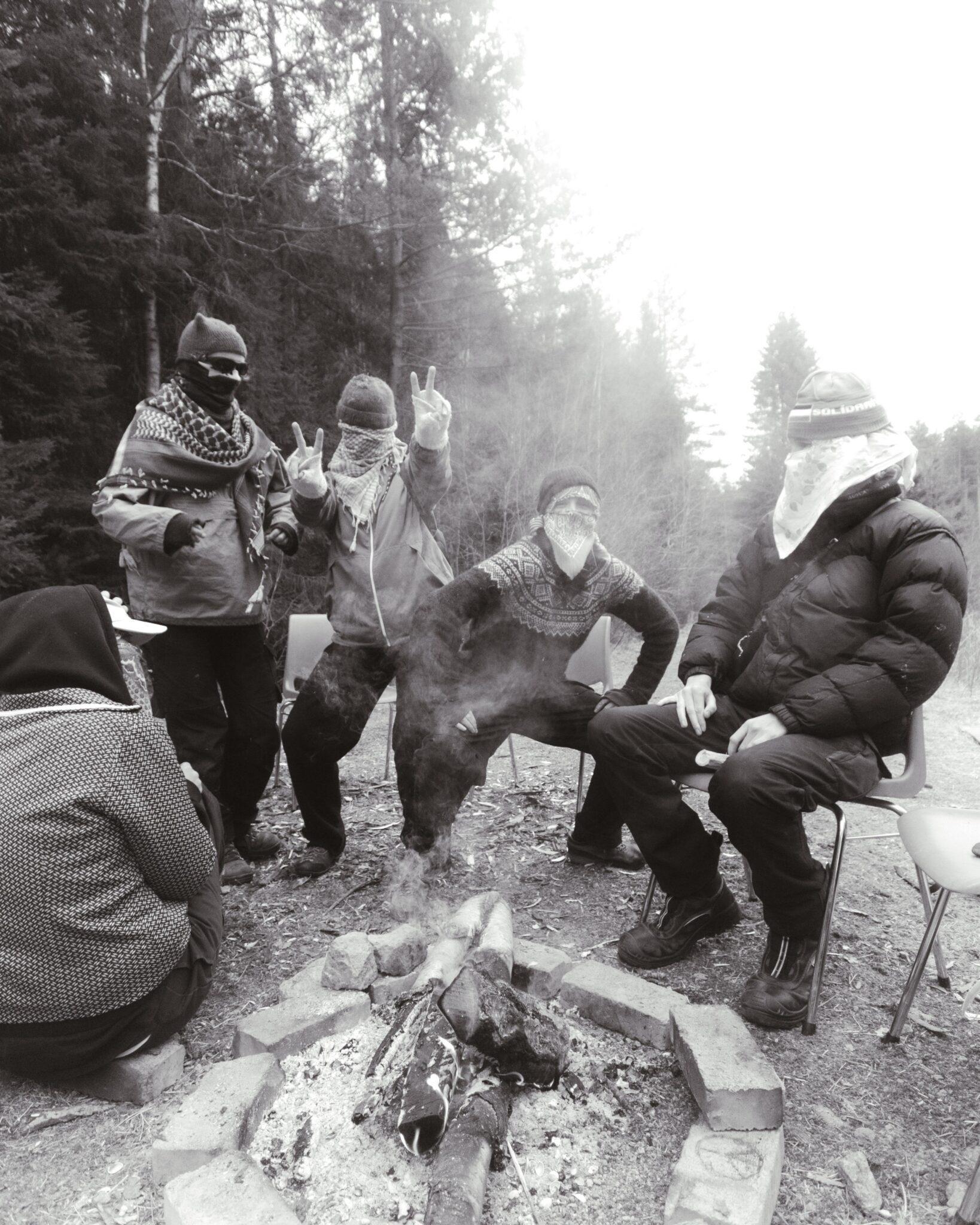 Five persons doing poses around a campfire. They are masked.
