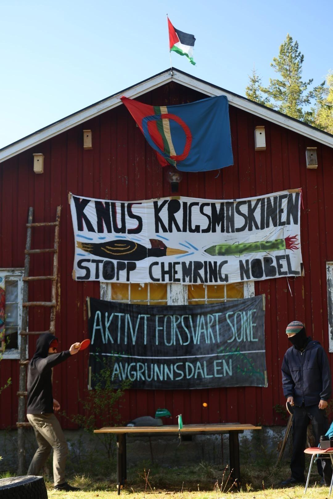 The front facade of the squatted house. In front of the house, two masked people are playing pingpong. On the facade of the house hangs various flags and banners. Topmost is the palestinian flag. Right underneath is the flag of Sápmi, the indigenous people whose land overlaps parts of Norway, Sweden, Finland and Russia. Next is a banner with the text "knus krigsmaskinen, stopp Chemring Nobel", and an illustration of a local bird flying directly against a missile. Lowest hangs a black banner with white text "aktivt forsvart sone - Avgrunnsdalen"