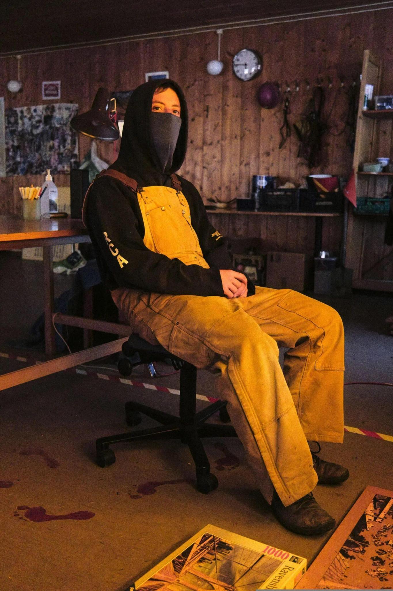 A photo inside the squatted house. A full body portrait of a masked person sitting in a chair. The person is wearing yellow overalls. On the floor is a 1000 piece puzzle.