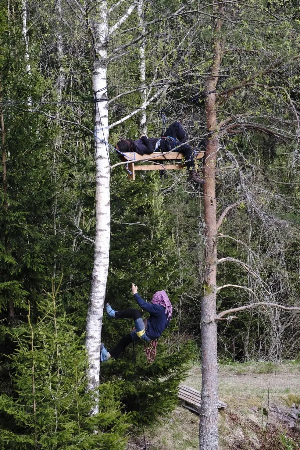 A diverse forest. High up between two trees is suspended a wooden bench. On the bench, a masked person is laying down. Another masked person is climbing up to the bench, supported by climbing rope.