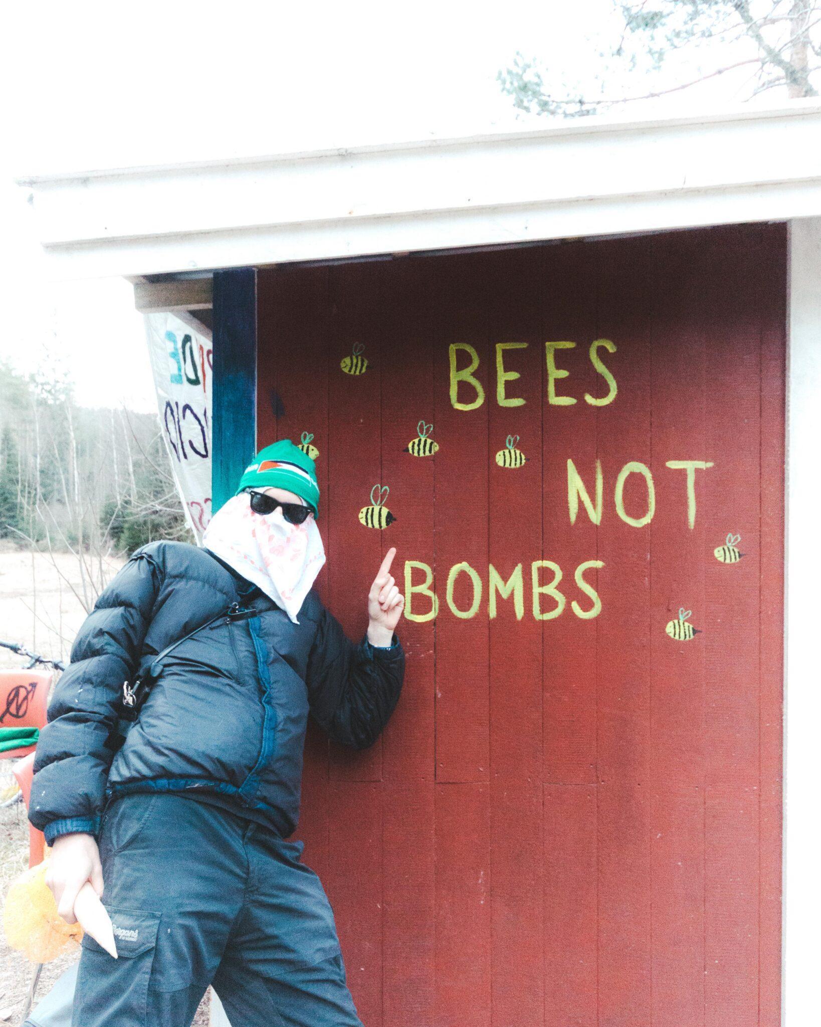 Photo of the info point, a movable shack used as an organized point of contact between the squatters and the rest of the public.

On the red wall is painted the yellow text "bees not bombs", and many bees are painted around it. A masked person is standing, leaning against the wall, and pointing at the text