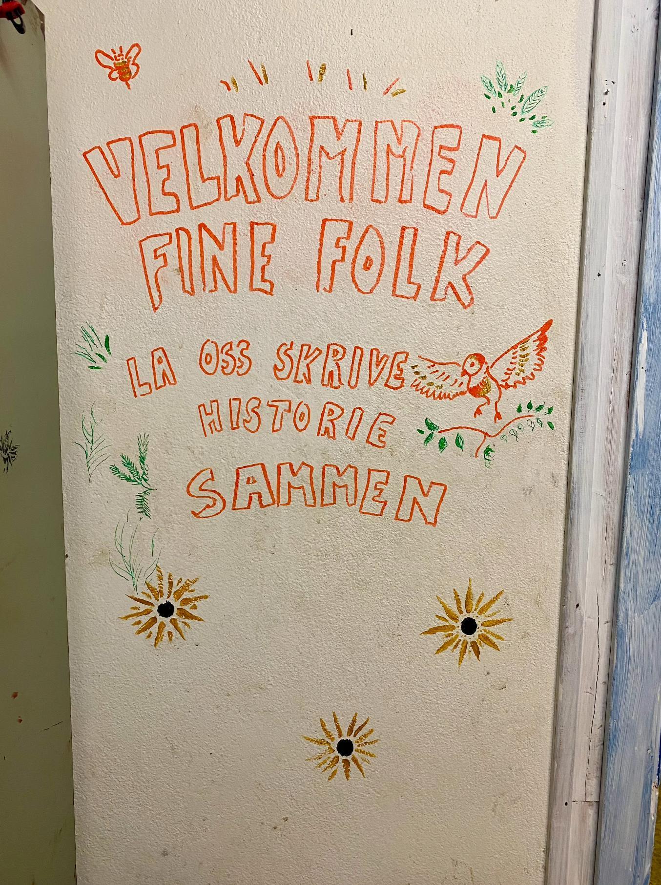 A white wall painted with the text "velkommen fine folk! La oss skrive historie sammen". Around the text is painted flowers, birds, and insects.