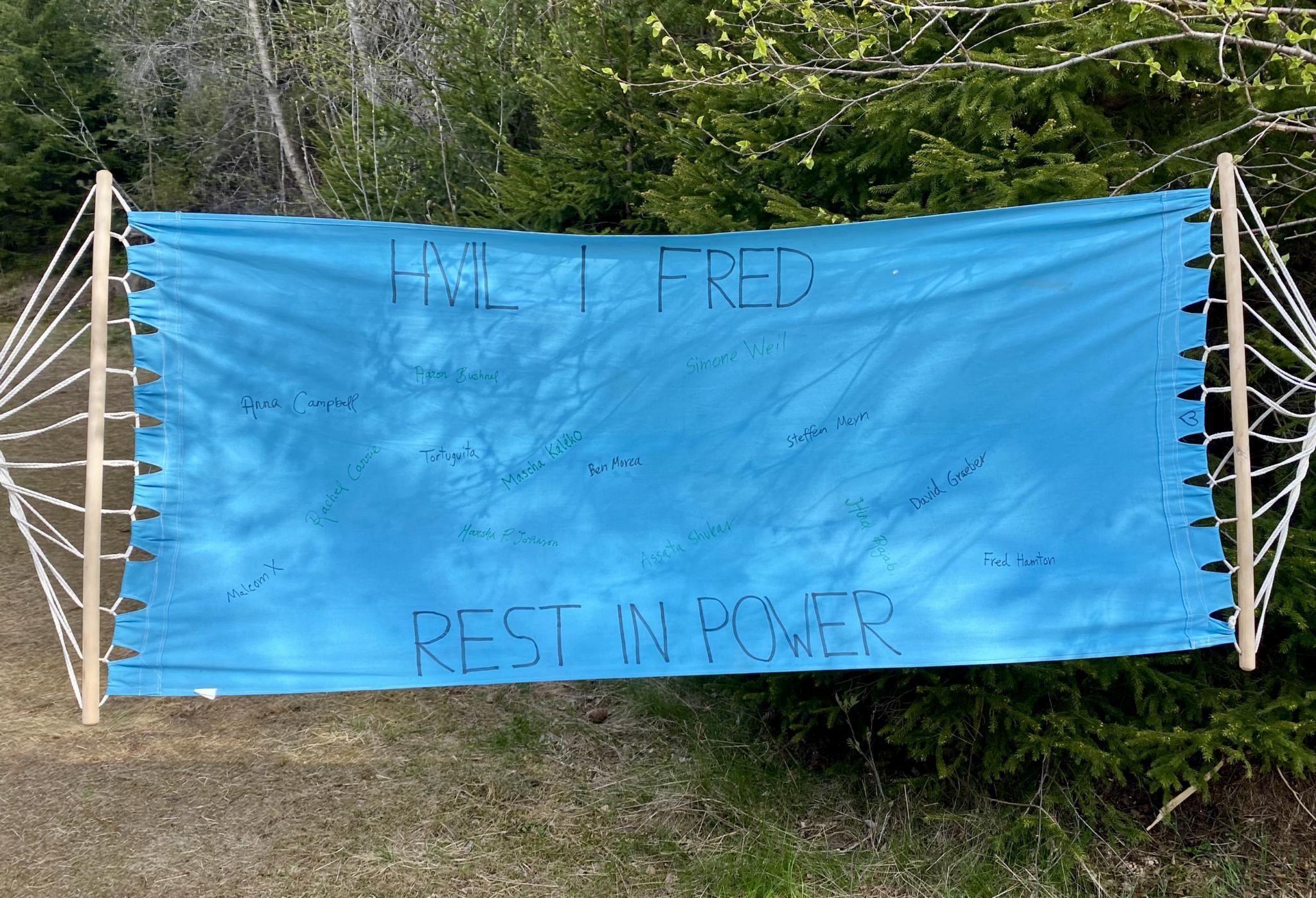 A light blue hammock. On the sheet is written the large text "hvil i fred, rest in power". In small text spread around on the sheet are written names of comrades who struggled for resistance, for liberation. Malcolm X, Anna Campbell, Marsha P. Johnson, Ben Morea, David Graeber, Fred Hampton, and many others