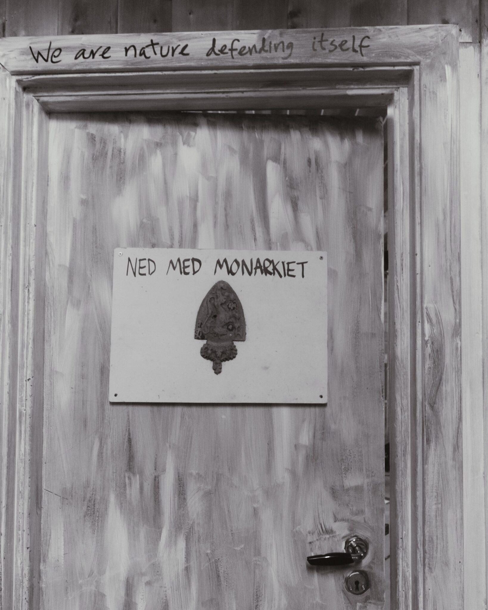 A greyscale image of a doorframe with a barely-open door. On the door frame is written the text "we are nature defending itself". On the door is mounted a plaque with the text "ned med monarkiet" and an upside-down symbol of the norwegian monarchy.