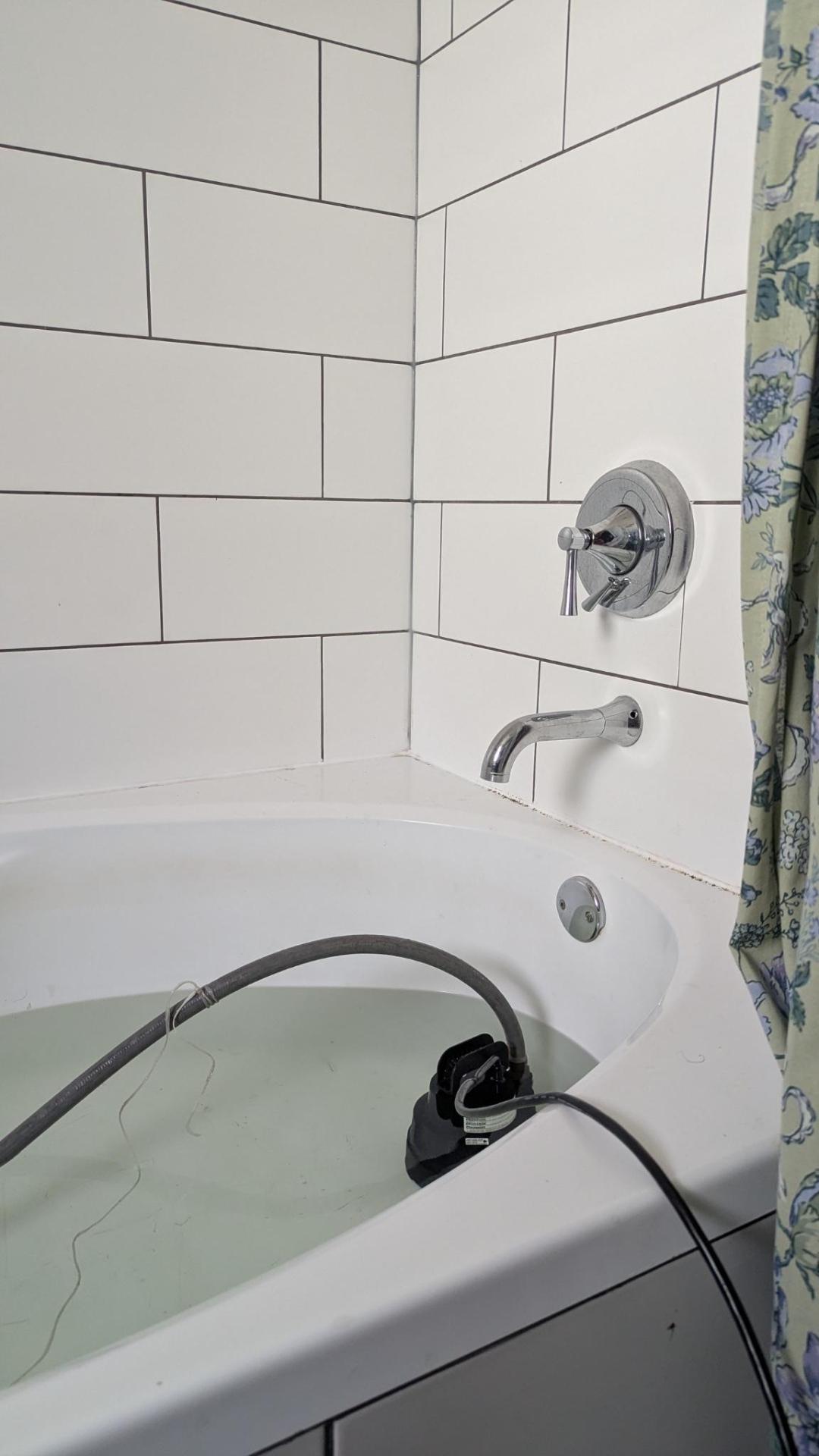 Photo of a white bathtub containing some water and a black pump about the size of a 1kg bag of sugar. A grey garden hose leads from the pump out of the frame, as does a black power cord. There is a string tied to the garden hose. A green floral cotton shower curtain is just visible on the right side