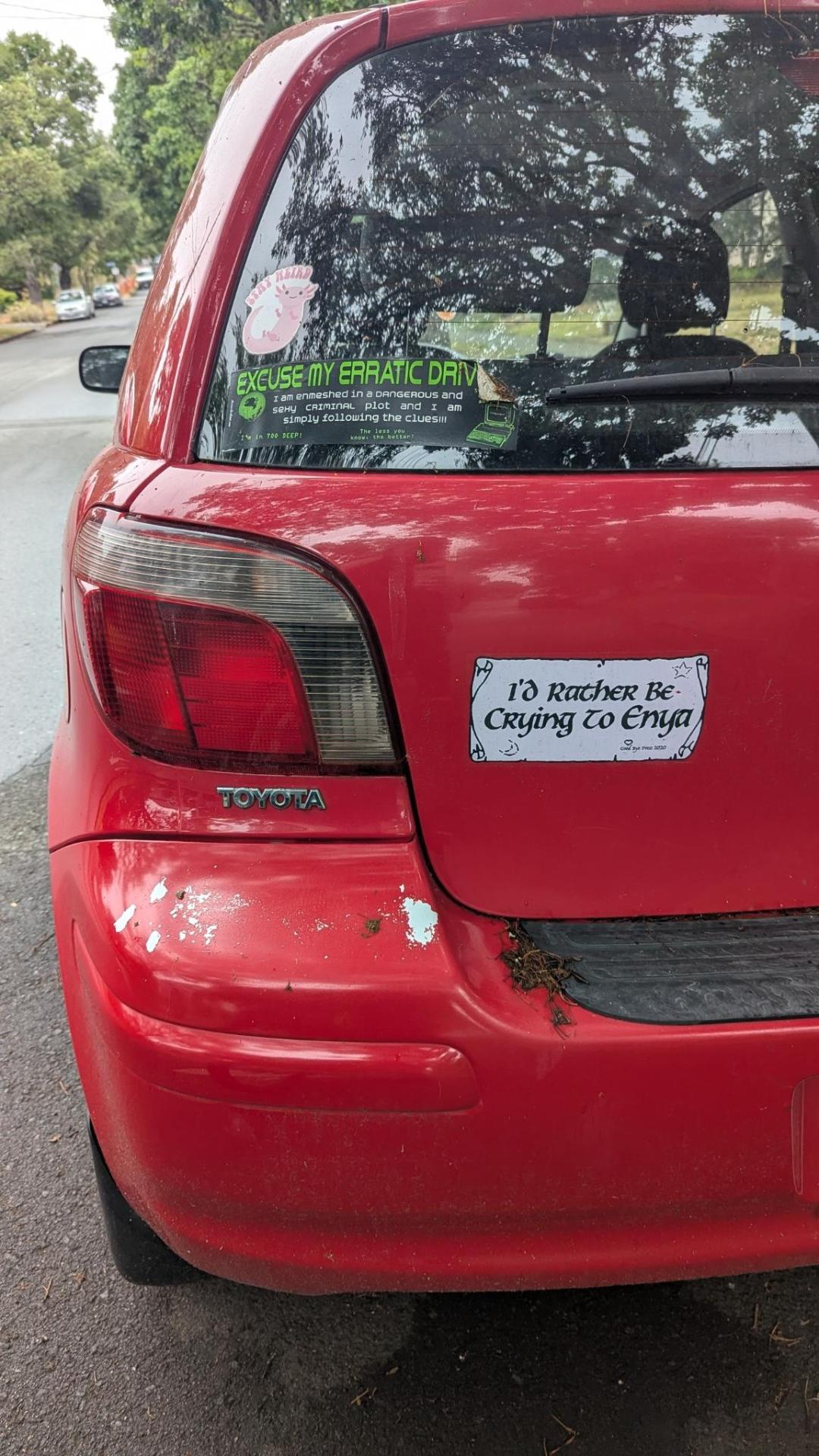 Photo of a bumper sticker on the back of a small red car. In uncial calligraphy it says "I'd rather be crying to Enya"