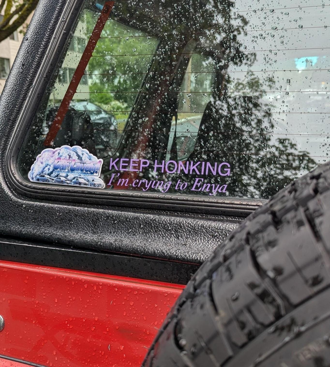 Photo of a transparent bumper sticker on the back window of a different small red car. Pink font says "KEEP HONKING I'm crying to Enya"