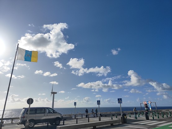 Vista del mar en una zona de aparcamiento, con el cielo azul y unas pocas nubes, y la bandera de Canarias ondeando