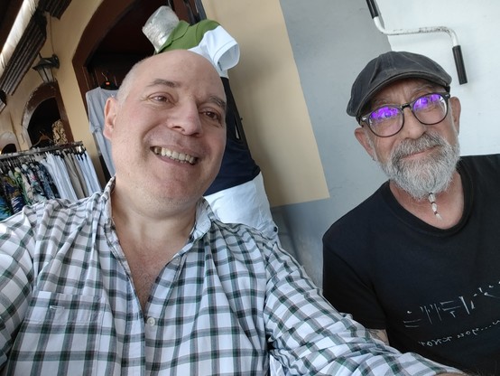 Selfi de Eleder, con camisa a cuadros desabrochada, y Loubam, con camiseta de Skyrim, gorra y elegante barbita blanca, sonrientes y a las puertas de un bar