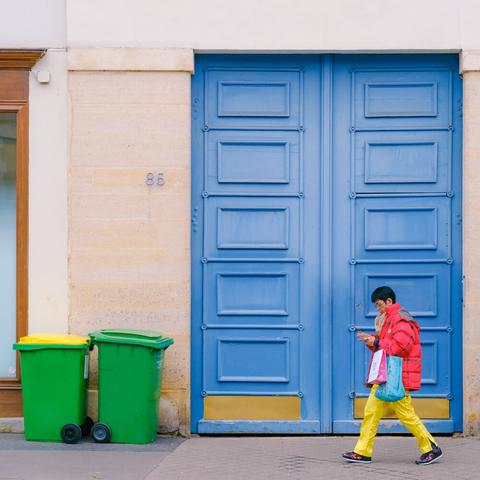 A woman in a red jacket and yellow pants is walking in front of a large blue door. Green trash bins are on the left side. One trash bin has a yellow lid.