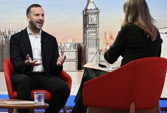 A TV Studio set up with Big Ben in Background; a man in bkack suit and white shirt, a woman in black top seen from back; both on red chairs, man talking, woman interviewing him. It’s Zack Polanski on BBC Laura Kuenssberg