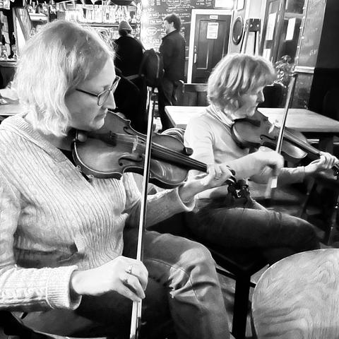 Two women side by side playing fiddle in a pub.