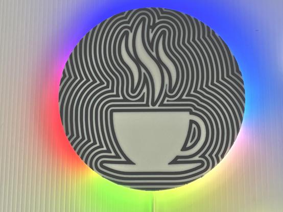The photo shows a circular HALO CLOCK featuring a stylized illustration of a steaming cup of coffee. The cup is white with a simple handle, and it's placed on a saucer. The background consists of bold, wavy black lines creating an optical illusion effect, with the steam rising from the cup integrated into the pattern. The clock is backlit, giving it a rainbow effect against a ridged, light-coloured surface.