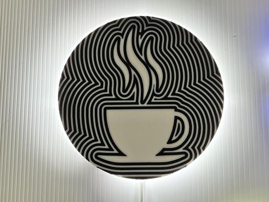 The photo shows a circular HALO CLOCK featuring a stylized illustration of a steaming cup of coffee. The cup is white with a simple handle, and it's placed on a saucer. The background consists of bold, wavy black lines creating an optical illusion effect, with the steam rising from the cup integrated into the pattern. The clock is backlit, giving it a glowing effect against a ridged, light-coloured surface.