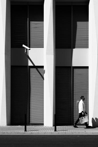 A monochrome photo of a person in a white shirt and black pants holding a bag in front of a building with closed blinds. A security camera is pointing at the person.