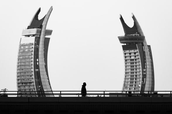 A silhouette of a person walking on a bridge. Two arched buildings in the background frame the subject.