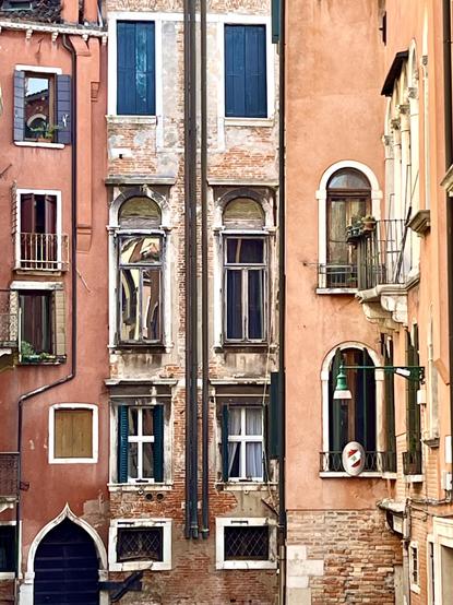 Colour photo of an exterior house wall, divided into three separate buildings - the left one painted a soft warm red, the middle one made of faded brick, and the one on the right painted a powdery orange - and what I would describe as an abundance, possibly even an excess, of windows, fifteen in all, of all different shapes and sizes. Some windows are rectangular, some square, some horizontal rectangles, some arched; a couple at the top have blue shutters, one has a little window box, one a miniature Juliette balcony; none of them have straight edges, and very few of them are in line with each other. The result is slightly disorientating, like a mad magic glazier has just thrown a bunch of excess stock at the wall and let them land where they may. It feels very emblematic of Venice in its beautiful chaos.