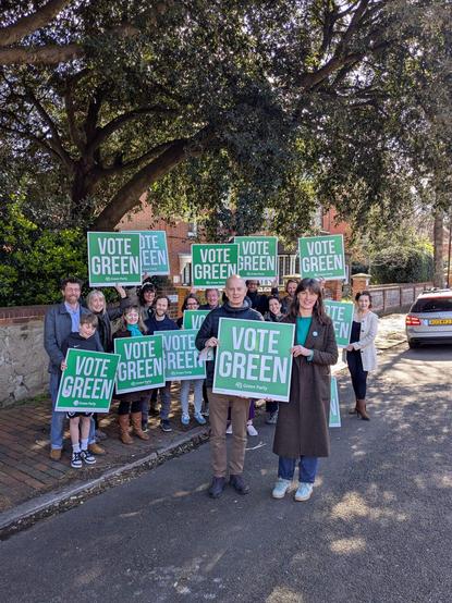 A group of 15 or so people on a road and pavement large trees behind, holding “Green Party” placards. Nice sunshine