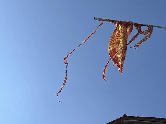 Colour photo looking up at a muted but clear blue sky, with a tiny sliver of a roof at the edge of the lower right corner of the frame. A horizontal flag pole emerges from the right hand side into the centre of the image, from which is hung the banner of San Marco: a golden lion on a red background, with six wide fringes on the flying-end of the flag. However, this particular specimen has clearly seen the harsh effects of some ferocious wind, and the flag has ripped almost all the way along the lines of the fringes, and then been blown and spun round and round the flagpole, so that what remains is a dense scrunched lump of fabric in the middle where the sun glints off the barely-visible wings of the lion, with long tattered ribbons on either side of it, which are dancing in the current breeze.
