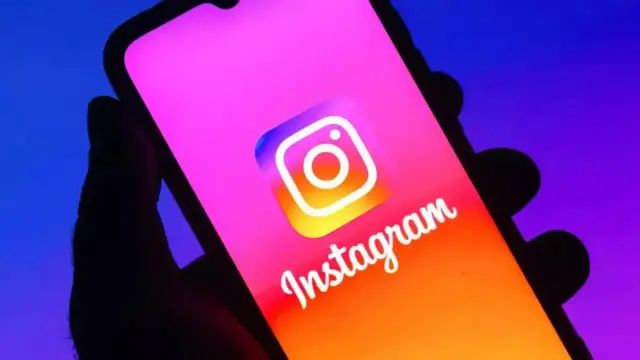 A hand holds a smartphone displaying the colorful Instagram logo against a gradient purple and blue background.